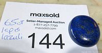 Close-up photo of deep blue lapis lazuli cabochon showing color and gold flecks on white maxsold auction card with lot number 144.