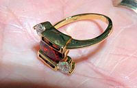 Close-up image of a gold-tone ring with a rectangular red man-made garnet stone centered and two clear round accent stones on either side. The ring is size 8.