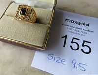 Gold tone men's size 9.5 ring displayed in box with auction lot number, showing black center stone and surrounding clear stones.
