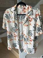 Front view of lightweight women's silk Hawaiian shirt with orange and gray floral pattern on cream background, hanging on black hanger.