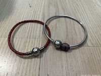 Two Pandora bracelets placed together on wood floor: a silver bangle with a pink heart charm and a red woven leather bracelet with silver clasp.