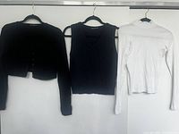 Black cropped cardigan, navy blue braided vest, and white turtleneck hanging on black hangers against white wall.