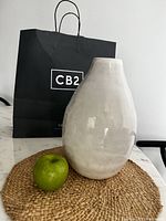 Front view showing the full glossy white vase on a woven placemat next to a green apple, with a black CB2 shopping bag behind.