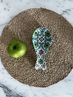 Photo showing top view of the ceramic spoon rest next to a green apple on a woven placemat on a marble surface.