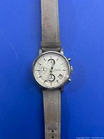 Top view of the Fossil silver wristwatch showing the white dial, chronograph subdials, date display, and worn grey leather strap laid flat on a blue background.