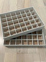 Two beige stackable jewelry organizer trays with multiple compartments for small jewelry items, stacked inside one another on a wooden floor.