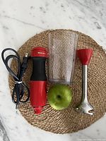 Red and black KitchenAid handheld mixer motor unit, detached stainless steel mixing blade attachment with red connector, clear mixing container, green apple for size reference on a woven placemat.