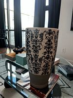 Full view of the vase showing the black floral damask pattern on crackled white glaze and unglazed lower portion on glass table with other home items in background.