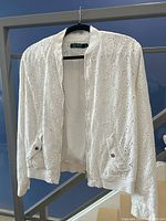 Full front view of the white eyelet patterned jacket hanging on a black hanger, showing overall design and ribbed collar, cuffs, and hem.