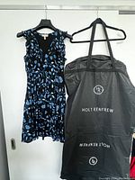 Black silk dress with blue floral pattern hanging next to a black Holt Renfrew garment bag, showing product presentation.