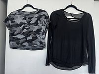 Both Lululemon workout shirts hanging on black hangers against a white wall, showing front views. One shirt is gray camo short sleeve with a front pocket, the other is solid black long sleeve with mesh upper sections.