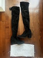 Full view of Stuart Weitzman black suede over-the-knee boots laid flat on wooden floor with dust bag underneath