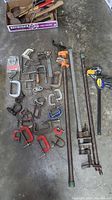 Overview of assorted clamps including C-clamps, pipe clamps, and quick-release clamps laid out on concrete floor.