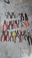 Top view showing a row of assorted pliers and cutters laid on a concrete floor, including red handled needle nose pliers, black handle cutters, orange handled wire cutters and aviation snips.