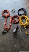 Photo shows six electrical cords (orange, yellow, black, white) coiled on concrete floor with various plugs and connectors.