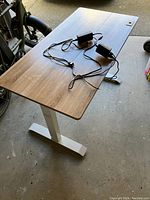 Full view of sit stand desk with wood grain rectangular top and white adjustable height legs, cords and power adapters on top.