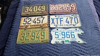 Overview of six vintage licence plates laid on dark quilted fabric, showing different years and designs from British Columbia and Northwest Territories.
