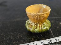 View of whole sunflower-shaped vase showing yellow scalloped top and green base on a black surface with a tape measure showing around 3 inches tall.