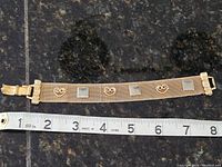Gold-tone metal mesh bracelet positioned next to measuring tape showing length approximately 7.5 inches, front side with decorative motifs visible.
