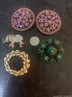 Top-down view of all five brooches arranged on a dark surface with a coin for size reference. Shows details of circular butterfly enamel brooches, rhinestone encrusted elephant, green floral and gold-tone leaf brooches.