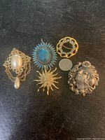 Top view of five brooches including a large pewter face brooch, a gold-tone pearl brooch with two pearls and chain, turquoise oval pendant brooch, gold-tone sunburst brooch and a blue rhinestone circle brooch. A quarter coin is placed for size reference.