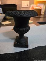 Front angled view of the vintage cast metal urn showing embossed details on the rim and middle section, with square base visible.