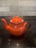 Front view of the orange enamel teapot on stove showing its round shape, orange body with brown handle and lid knob.