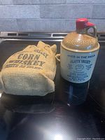 Stoneware whiskey bottle with red wax seal and embossed McCormick on top, next to burlap sack on a black glossy stove surface.