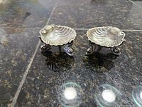 Pair of ornate silver salt cellars shaped like shells with dolphin feet on dark surface.