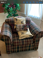 Plaid upholstered armchair set in a corner with three decorative pillows on the seat. The chair features rolled arms and has a skirted bottom. Surrounded by curtains and light from a nearby window.