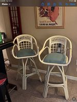 Two rattan bar stools with armrests and light blue cushions shown next to wall with artwork