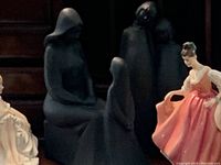 Five Royal Doulton figurines on a wooden surface; includes two large black matte finish figurines with abstract shapes and three detailed porcelain figurines in colorful dresses.
