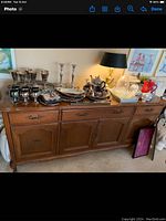 Photo shows a wooden dining room buffet with four drawers and cabinet doors. Various silver items, candlesticks, trays, a black and gold lamp, and decorative pieces are placed on top.