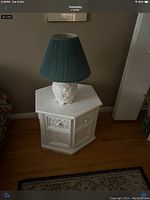 Photo of a white octagonal end table with decorative carved panels and a white ceramic table lamp featuring a floral relief base and teal pleated lampshade