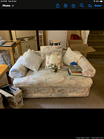 Front-facing photo of a floral patterned loveseat sofa with cushions. The sofa is pale with pink, blue, and green tones. There are five cushions on the sofa, including a small white lamp and books placed on it.