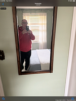 Photo showing full view of the rectangular mirror mounted on the wall, reflecting a man holding a phone and part of a bedroom with a bed and window with curtains.