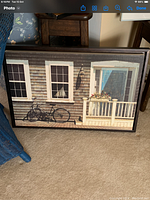 Framed picture showing the exterior of a house with wooden siding, two windows, a bicycle leaning against the wall, and a flower pot on the porch.