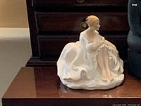 Porcelain figurine of seated woman in white dress holding floral garland across lap, positioned on wooden surface.