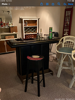 Photo of small home bar with black countertop, built-in wine rack and shelving, alongside black wooden stool with circular colorful seat on carpeted floor.