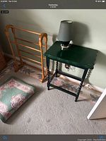 Photo showing a green wooden end table with a lamp on top, wooden valet stand, and floral cushion on carpeted floor.