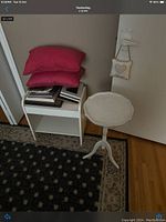 Photo showing small white wooden side table with scalloped top and matching tripod legs positioned against a white door, beside a small white shelf unit holding several books. Two red pillows are stacked on top of the shelf unit. A small fabric wall hanging featuring a heart design is attached near the door handle.