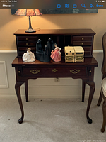 Photo showing small wooden secretary desk with decorative brass handles and curved legs. Several porcelain figurines and small storage containers are on top but not included in lot.