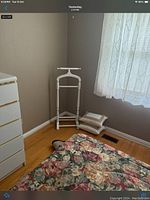 Wooden white valet stand placed near a wall and window with sheer curtain, next to a dresser and cushions on floor.