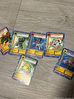 Photo showing 6 vintage Digimon cards fanned out on wooden surface. Cards show various characters with detailed stats and names.