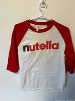 Full view of the Nutella long sleeve shirt with red sleeves and collar, white body, on a hanger.