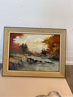 Frontal view of the framed landscape painting showing river, rocks, and fall foliage.