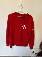 Front view of a red crewneck sweater with TFC logo on the chest hanging on a white hanger