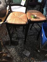 Two matching wooden bar stools with woven natural-colored rush seats and black wooden frames, placed on gravel surface.