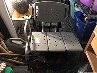 Photo of a gray plastic bathtub transfer bench with three sections, perforated seat, armrests, and adjustable aluminum legs in a cluttered indoor space.