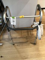 Side and front angled view of a silver aluminum adjustable medical walker on a wood floor with a plastic bag tied to one side.